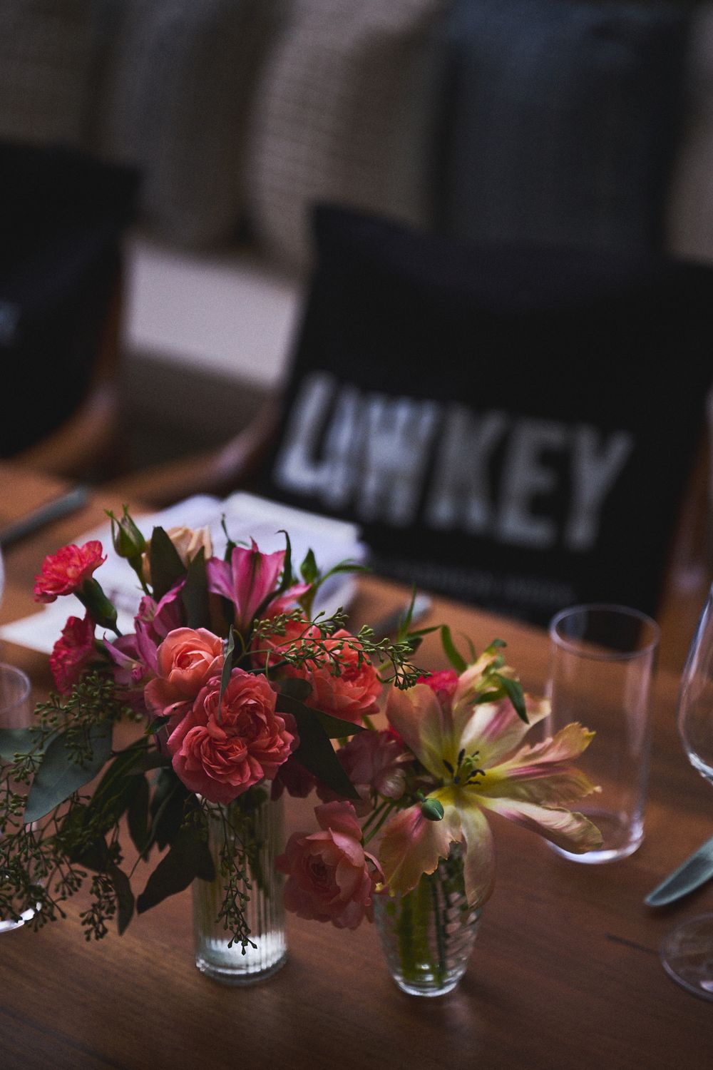 Small glass vases of coral roses and pink lilies on a wooden table beside empty glasses and a black pillow reading LOWKEY.