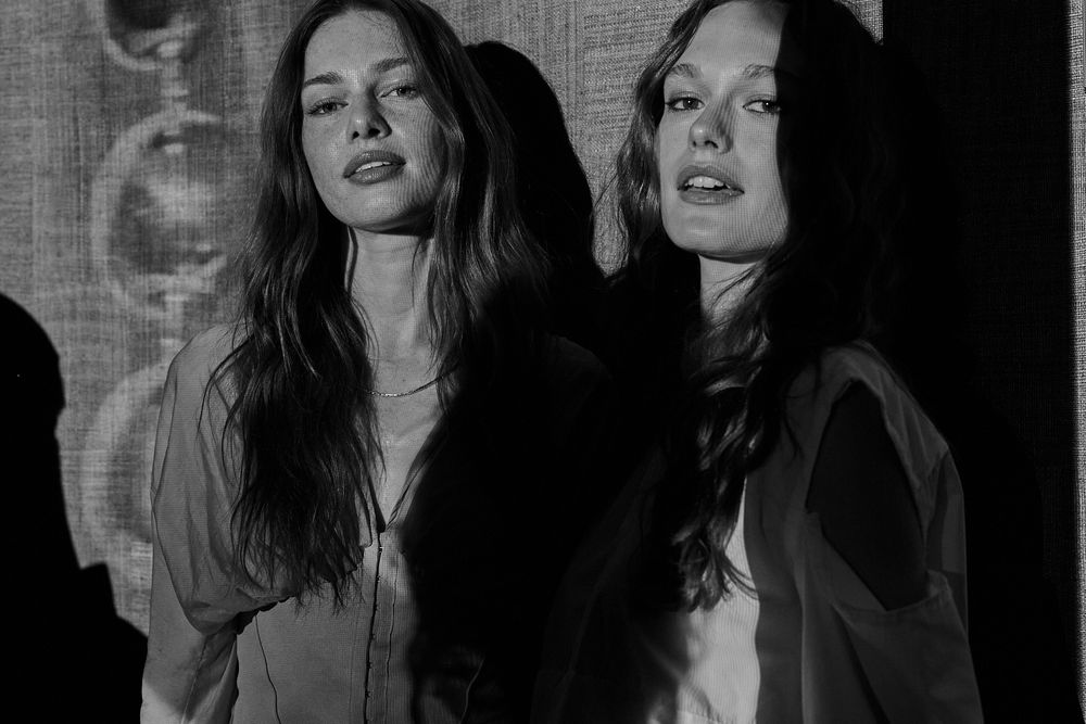 Black and white portrait of two women with long wavy hair posing side by side in moody, dramatic lighting against a textured backdrop.