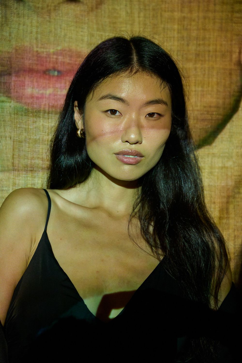 Woman with long dark hair and gold earrings wearing a black camisole, posed against a textured backdrop with a projected image of lips.