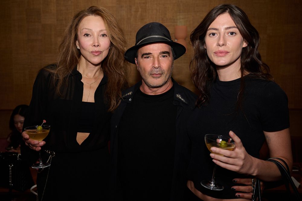 Three people in black outfits pose together in a dimly lit bar, two women holding cocktail glasses and a man in a black hat between them.