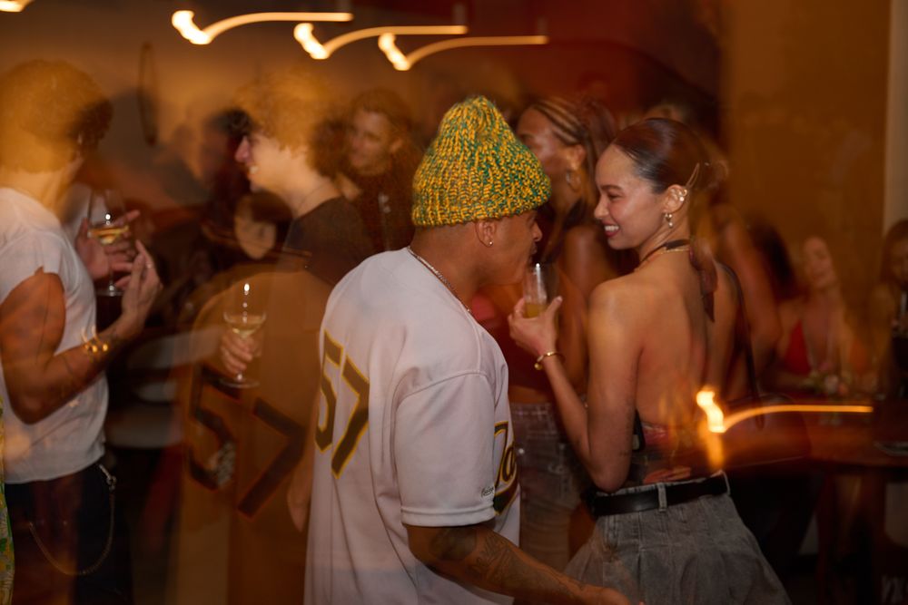 Man in a green knit beanie and white jersey chats with a smiling woman in a halter top at a lively, dimly lit party.