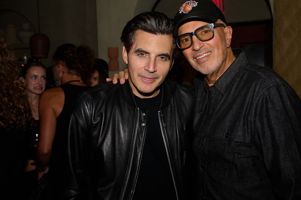 Two men pose closely at a lively indoor party, one in a black leather jacket and the other in a baseball cap and glasses, with guests behind