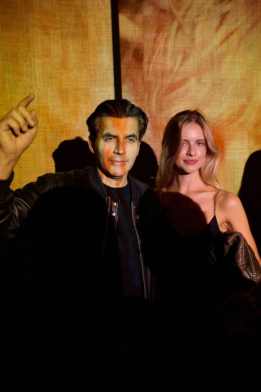 A man in a black jacket stands beside a smiling woman with long blonde hair as orange projected light plays across his face.