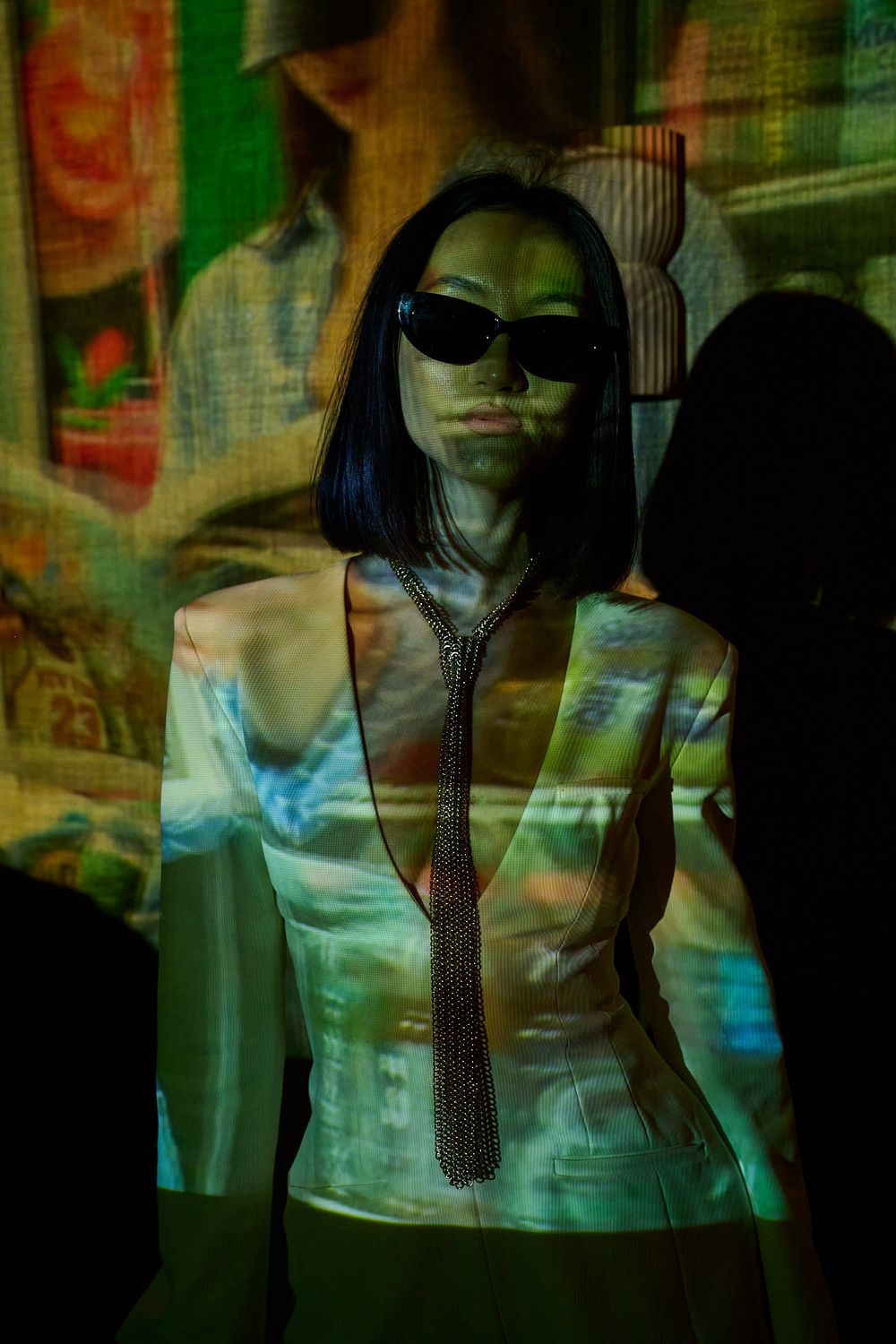 Woman with a bob haircut and dark sunglasses wearing a beaded necktie, illuminated by colorful projected imagery in a dark room.