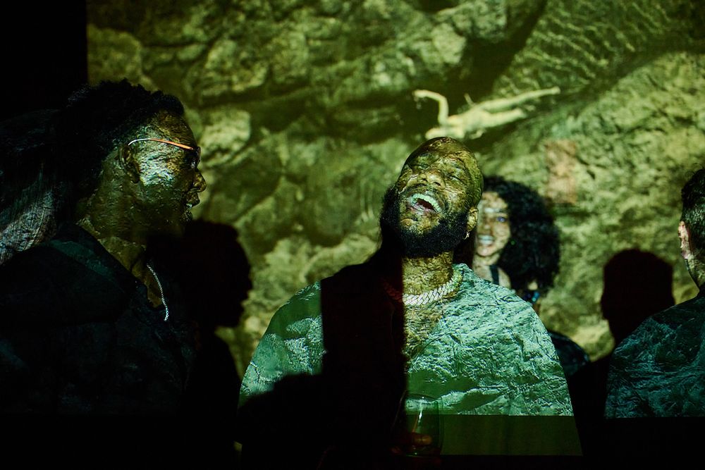 Group of people in dark clothing standing together in a dimly lit room with a green rocky cave-like wall projected behind them.