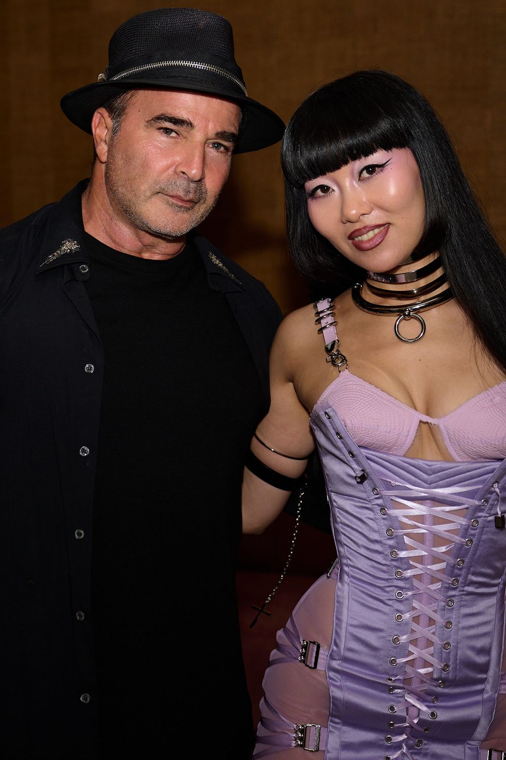 A man in a black hat and dark jacket poses beside a woman with black bangs wearing a lavender lace-up corset and layered choker necklaces.