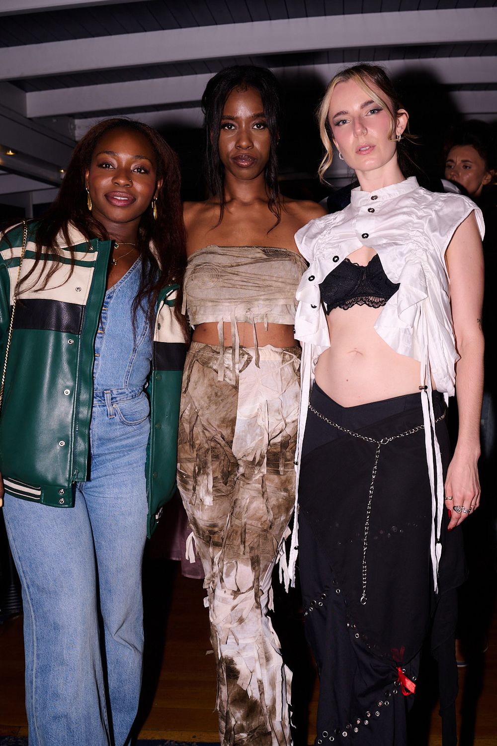 Three women pose together indoors in stylish outfits: a green varsity jacket with jeans, a printed strapless set, and a white crop top with 