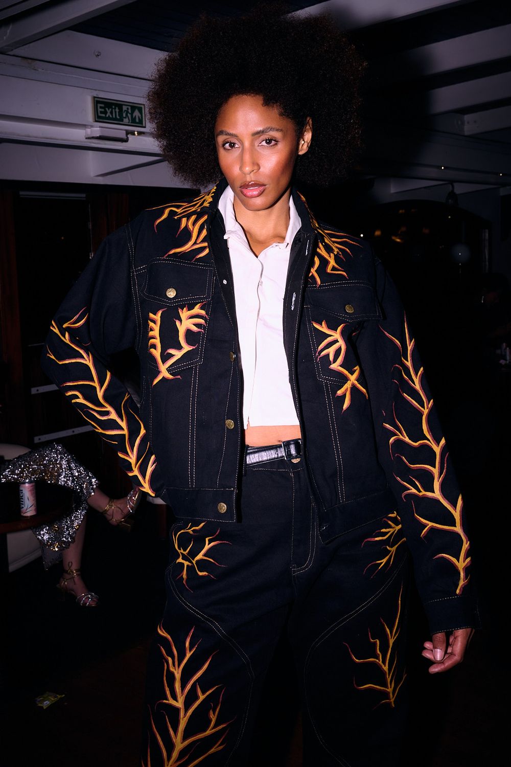 Model poses in a black denim jacket and matching pants with orange flame embroidery over a white collared top, with an exit sign behind.