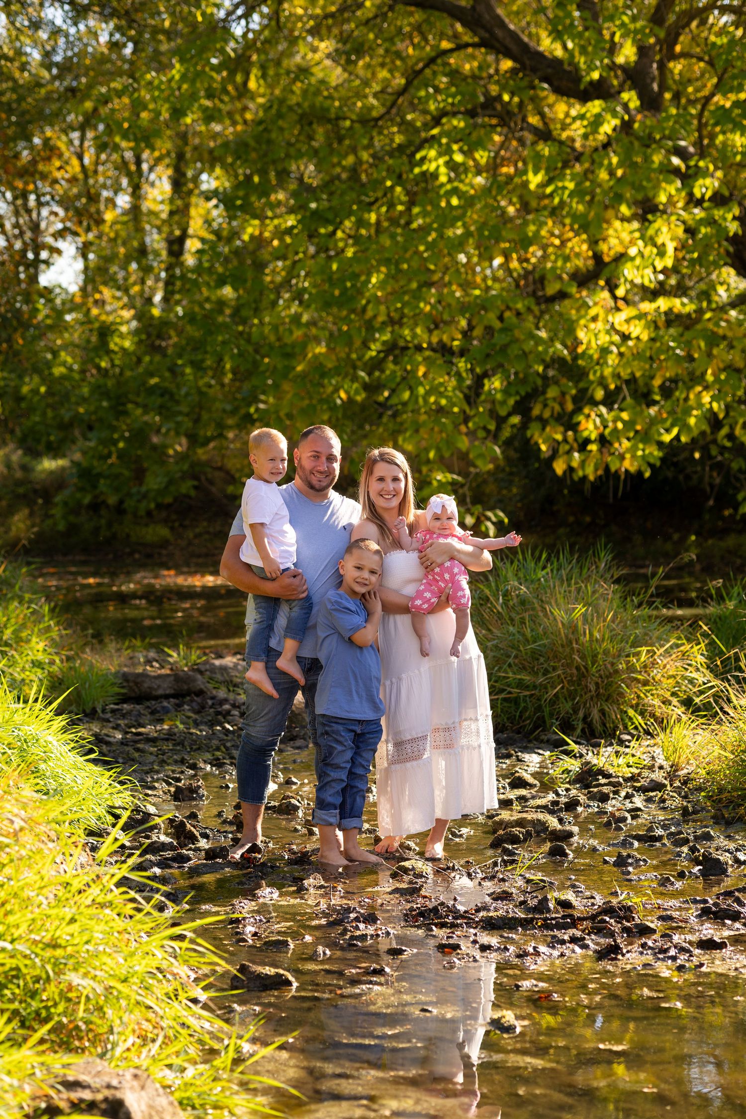 Happy family of five standing barefoot in a shallow creek surrounded by autumn foliage.