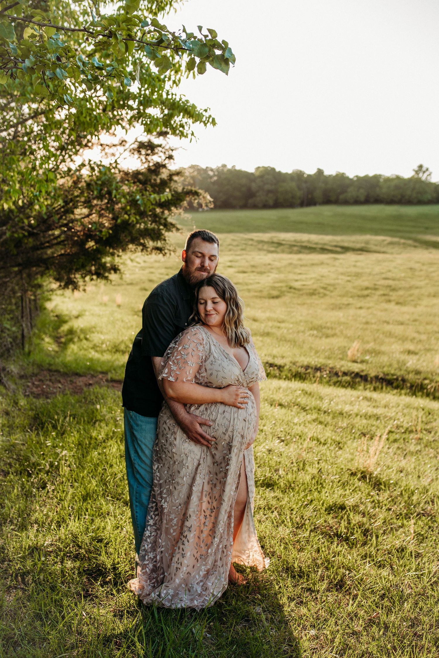 A couple poses in a sunlit field, the woman in a floral gown cradling her baby bump during a maternity photoshoot.