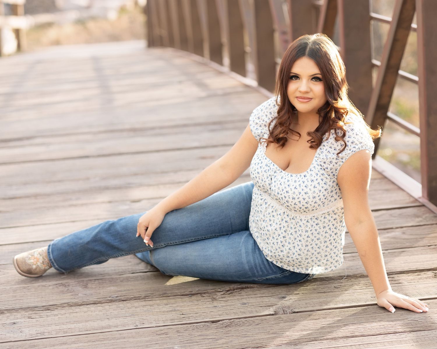 Young woman in jeans and white top sitting on wooden bridge, posing for senior portrait photo outdoors.