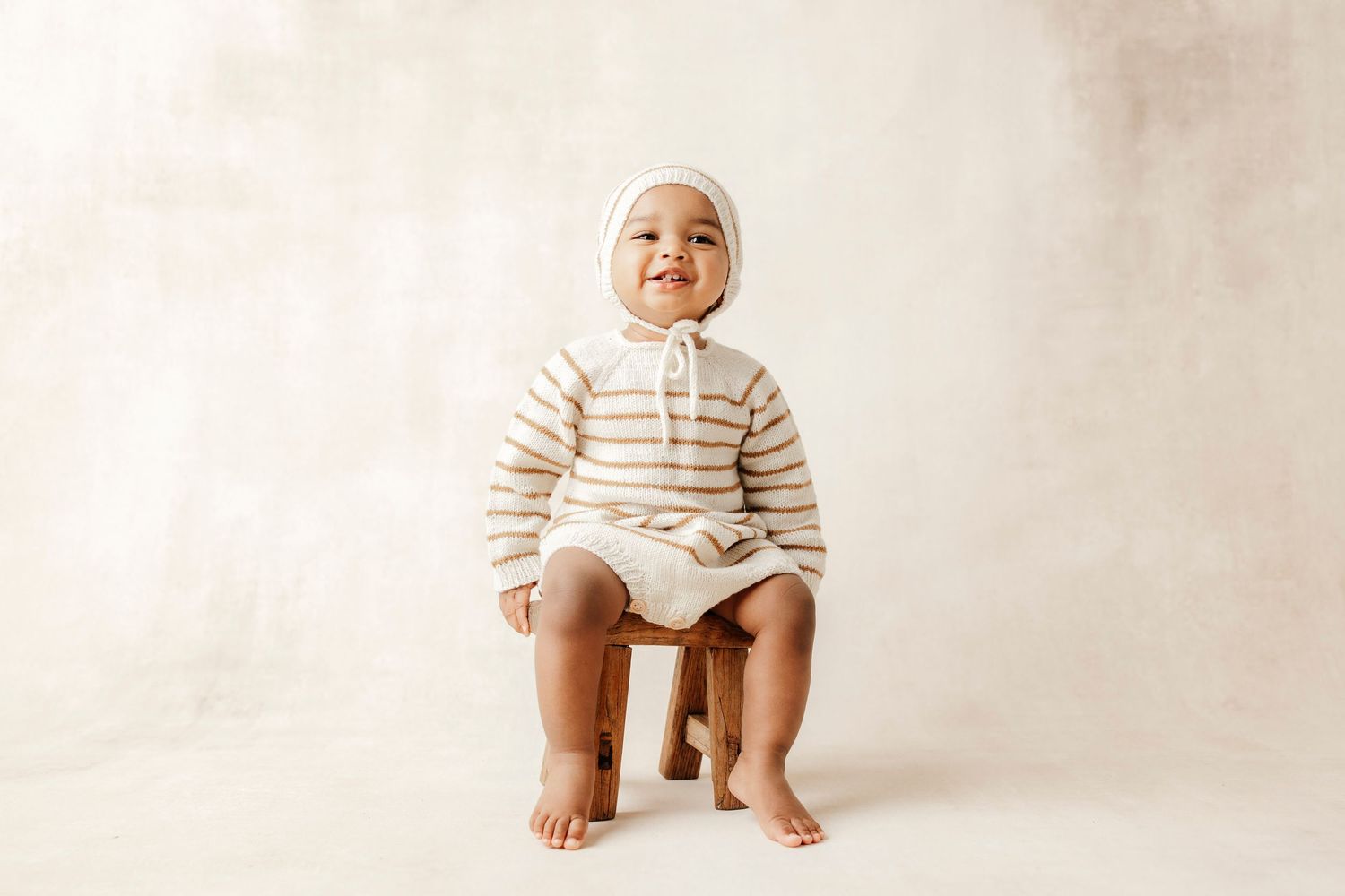 Smiling baby in striped knit sweater and bonnet sitting on a small wooden stool against a neutral background.