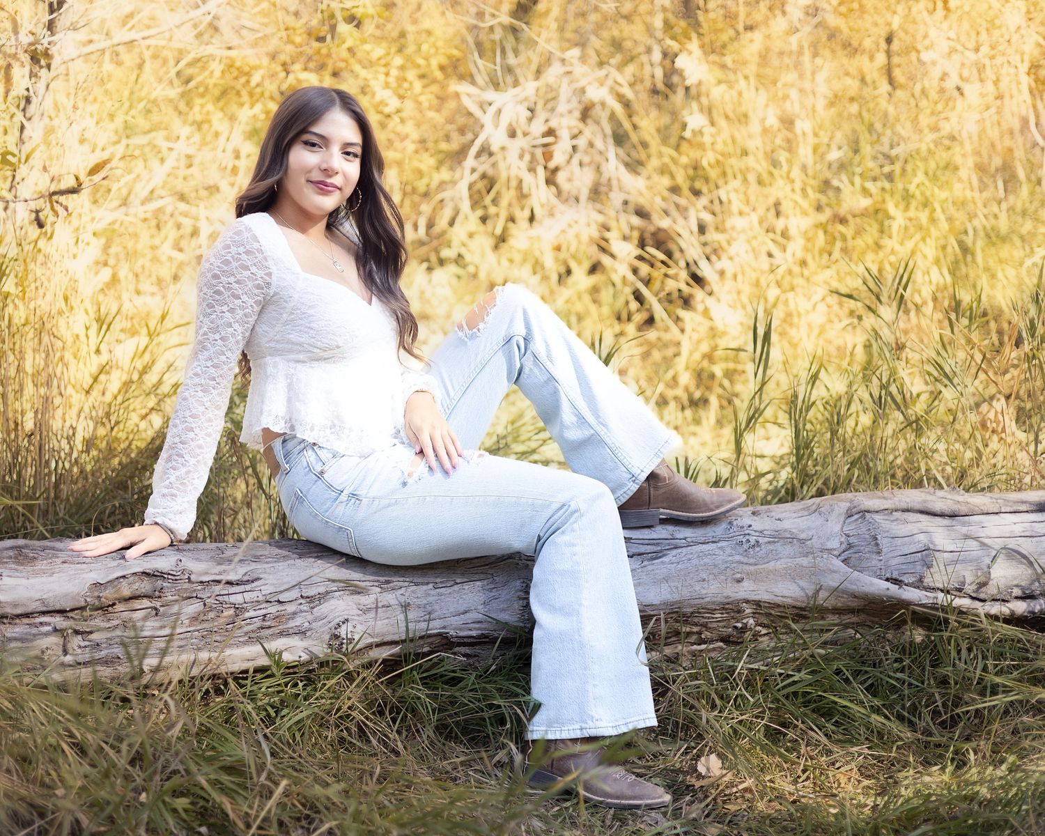 Young woman in white lace top and light jeans sitting on a log in a golden autumn field.