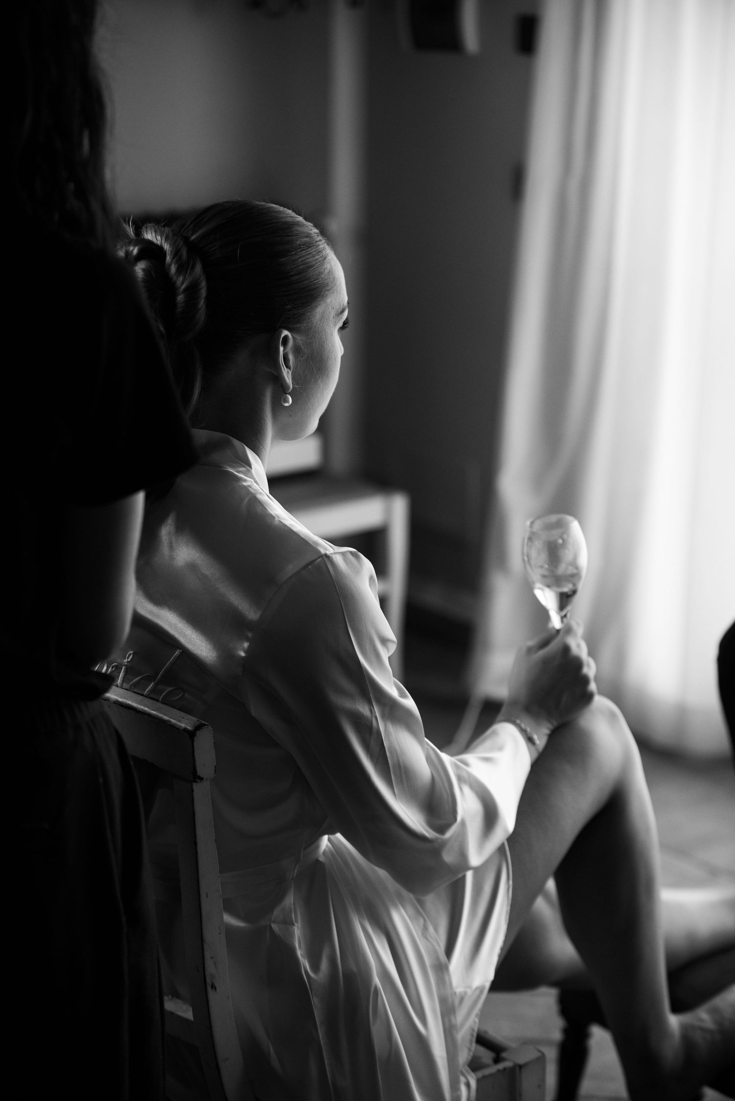 Black and white photo of elegant woman in silk robe holding champagne glass, seated by window in soft light.