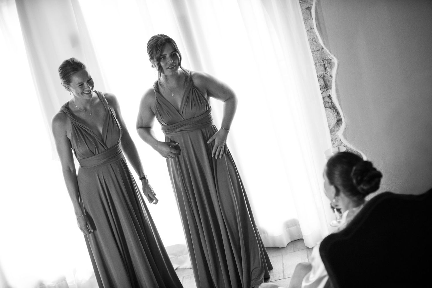 Two bridesmaids in elegant wrap dresses stand smiling in a bright room during wedding preparations.