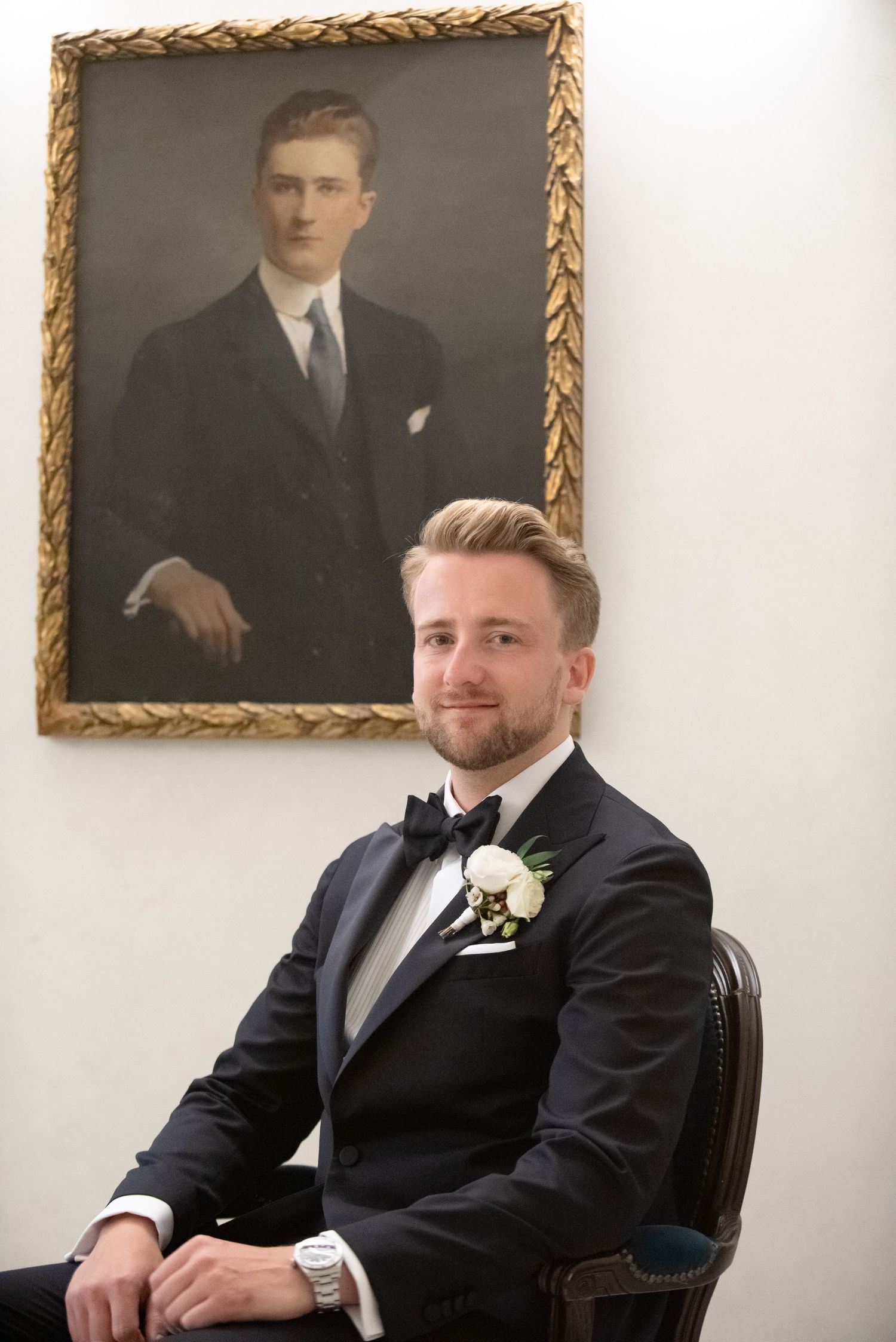 Groom in tuxedo with boutonniere sits before vintage portrait painting at wedding venue.