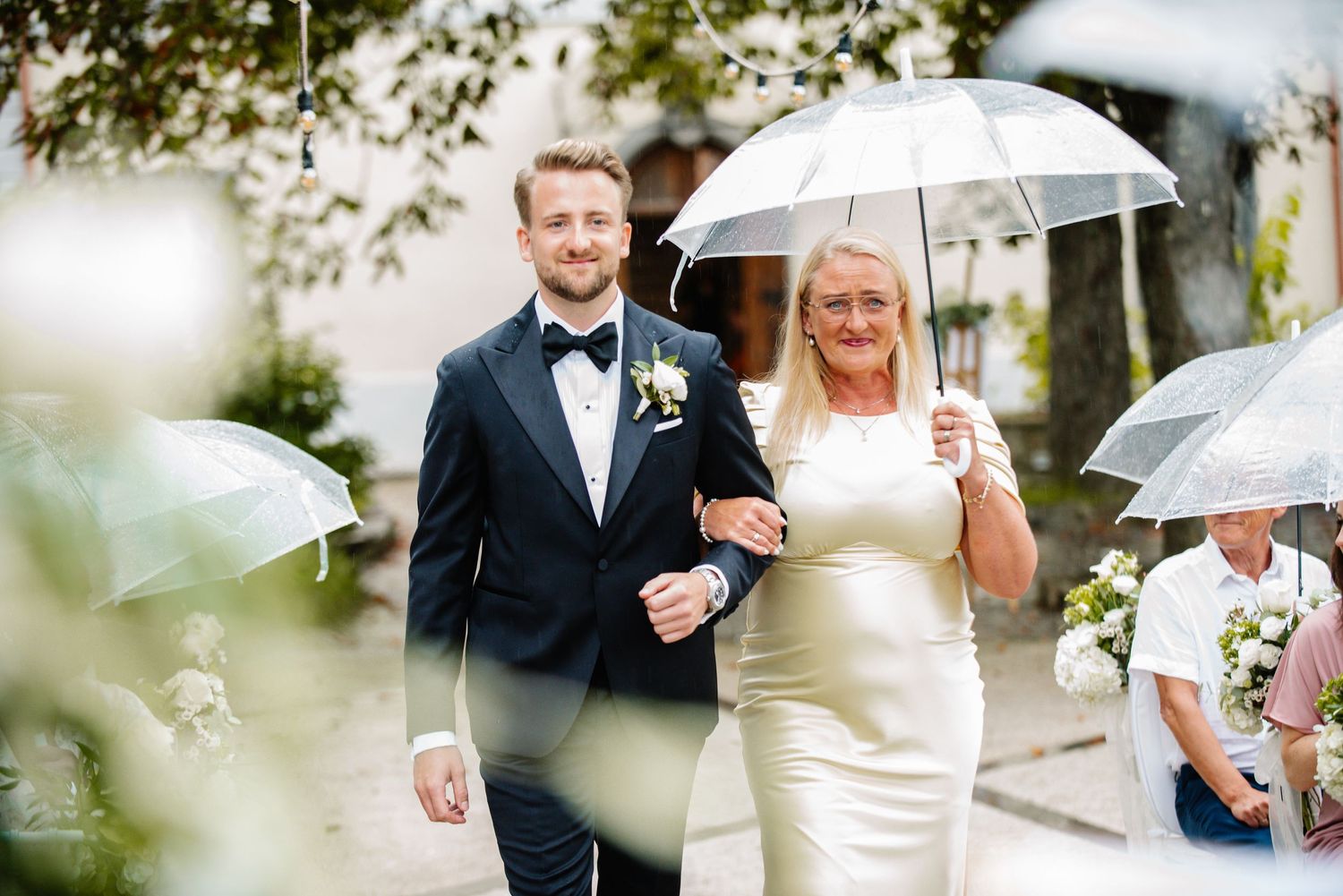 Groom in tuxedo walks with woman in white dress under clear umbrella at outdoor wedding ceremony.