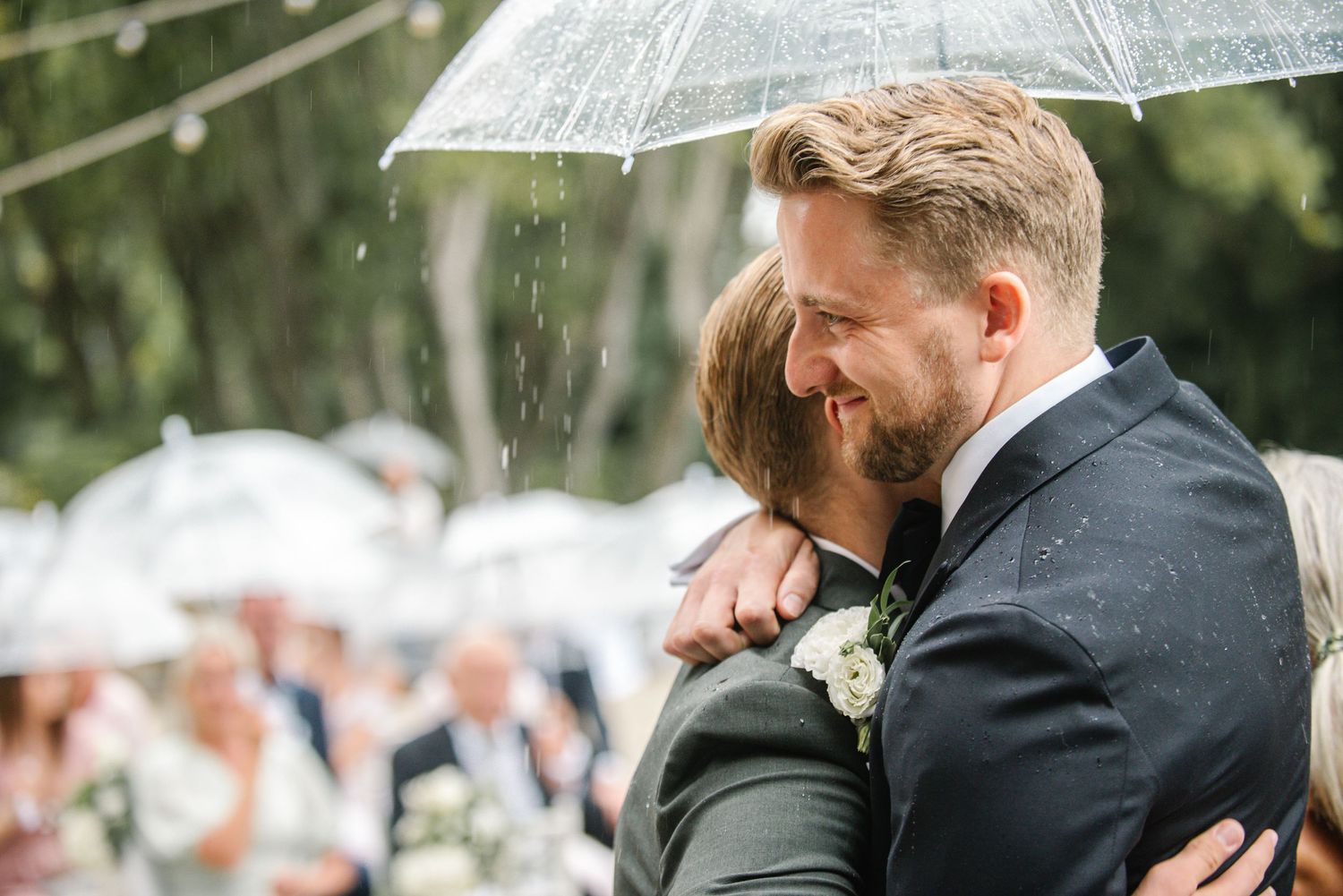 Couple embraces under clear umbrella in rain at outdoor wedding ceremony with guests in background.
