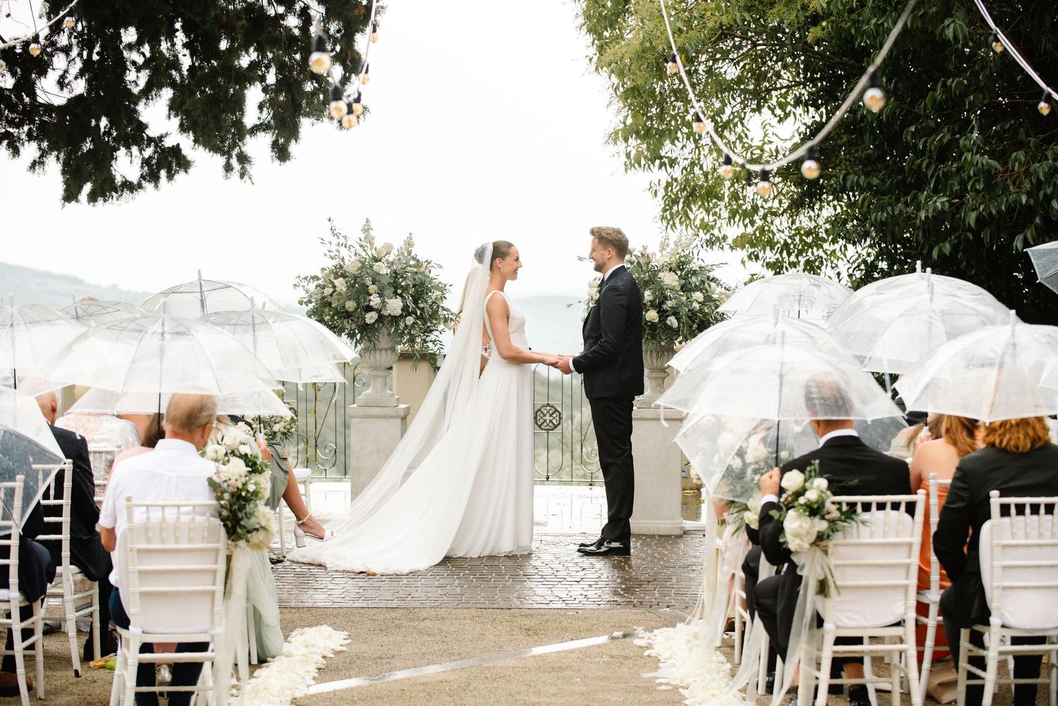 Outdoor wedding ceremony with bride and groom holding hands, guests with clear umbrellas seated on white chairs.