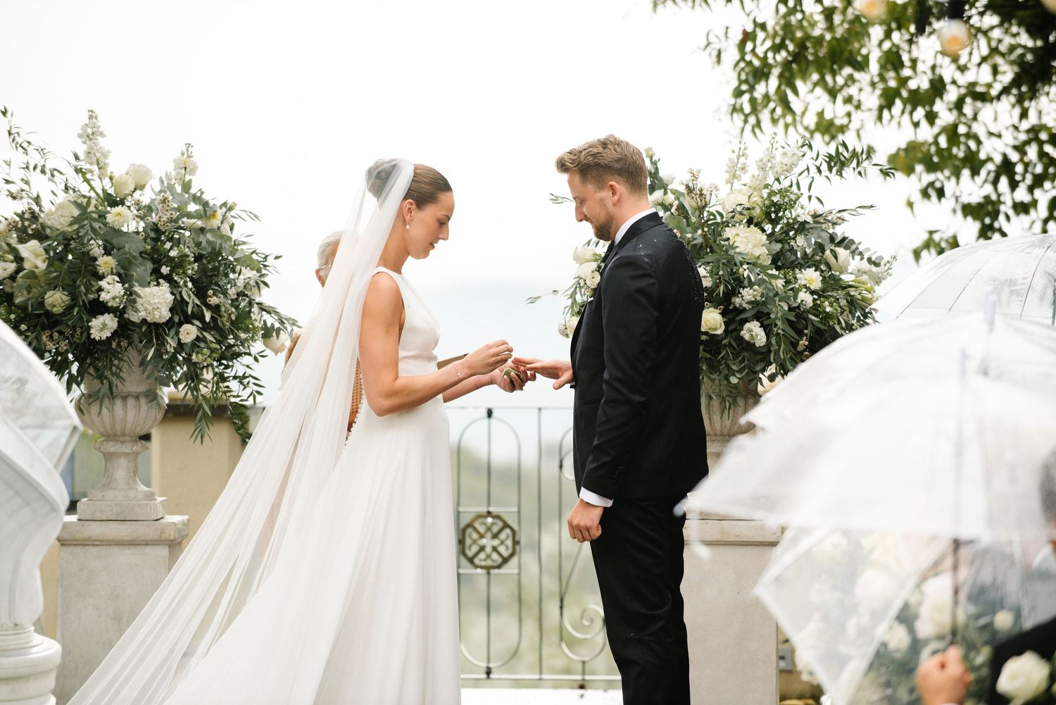 Bride and groom exchange rings at an elegant outdoor wedding ceremony with white floral arrangements and ocean views.