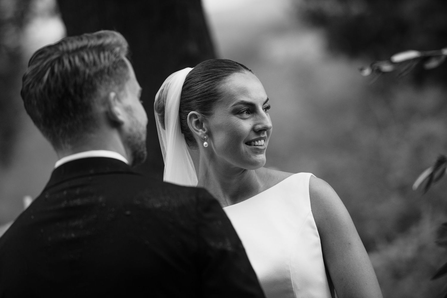 Black and white photo of a bride smiling at her groom during an outdoor wedding ceremony.