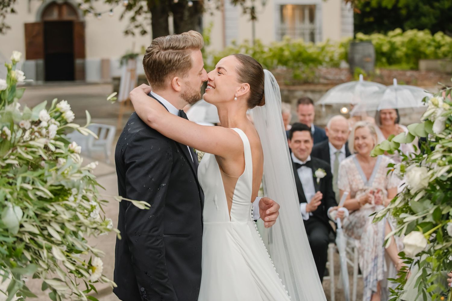 Bride and groom share a romantic kiss at outdoor wedding ceremony with guests applauding in background.