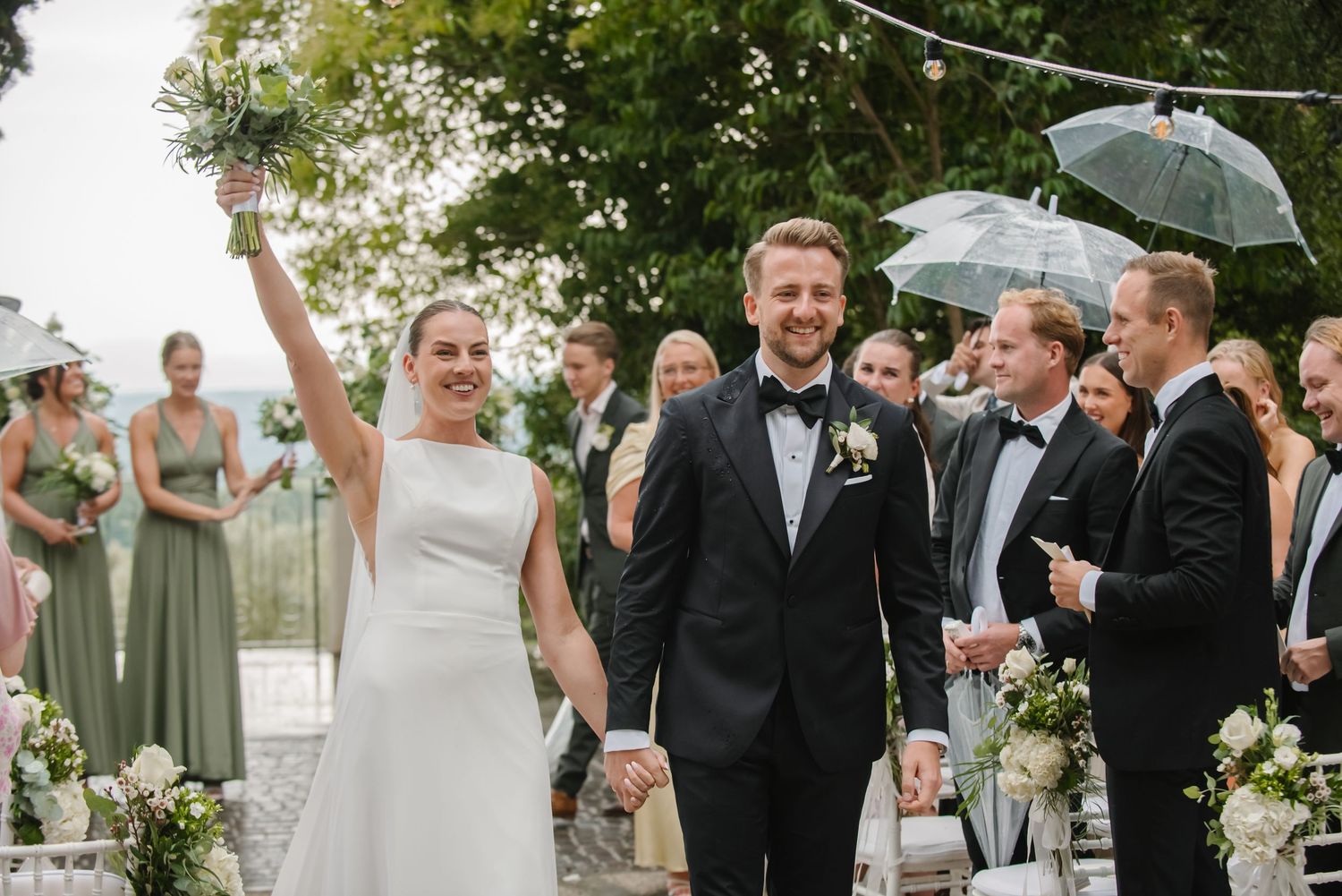Joyful bride and groom walk down the aisle after wedding ceremony, guests holding umbrellas and celebrating outdoors.