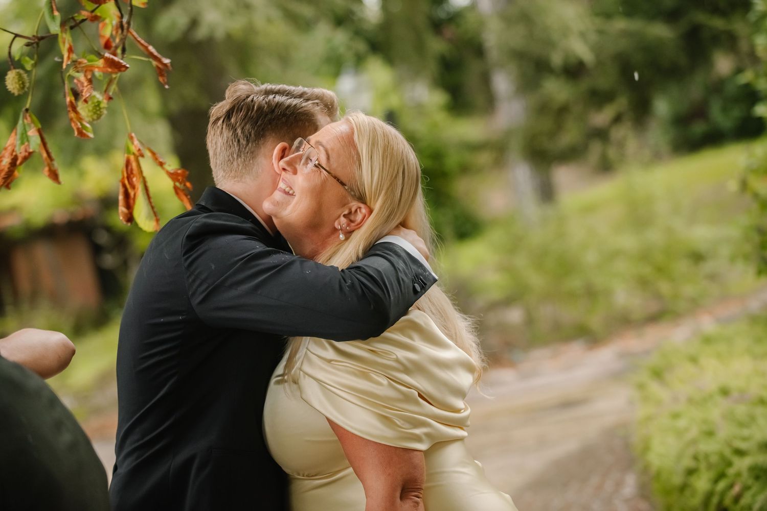 Couple sharing a romantic kiss outdoors, surrounded by lush green trees and red hanging flowers.