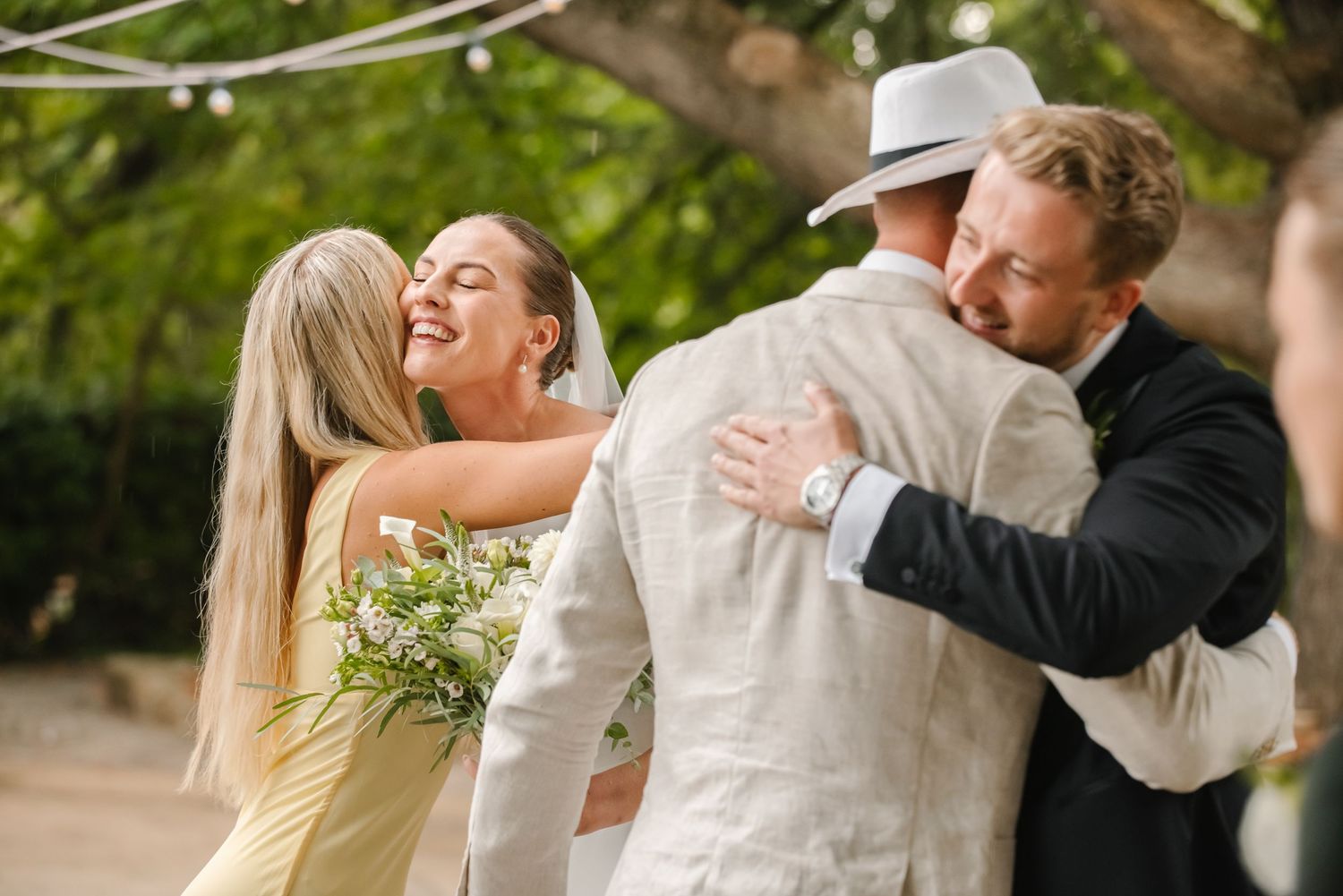 Bride and groom embracing wedding guests outdoors, smiling joyfully, surrounded by lush green trees at a garden ceremony.