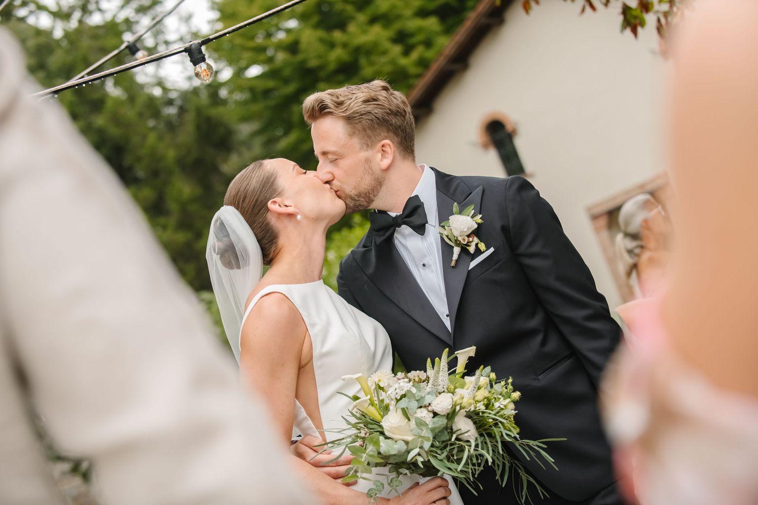Bride and groom share a kiss outdoors, bride holding a lush bouquet, groom in a tuxedo.