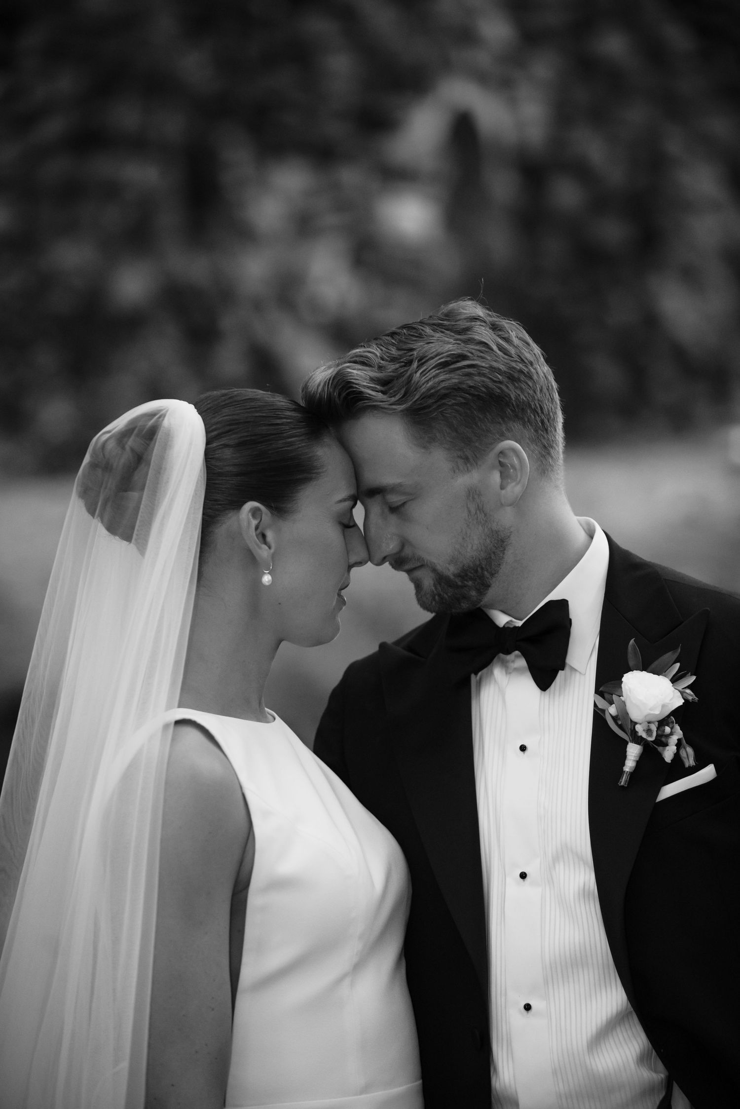 Black and white photo of bride and groom touching foreheads tenderly, bride in veil, groom in tuxedo with boutonniere.