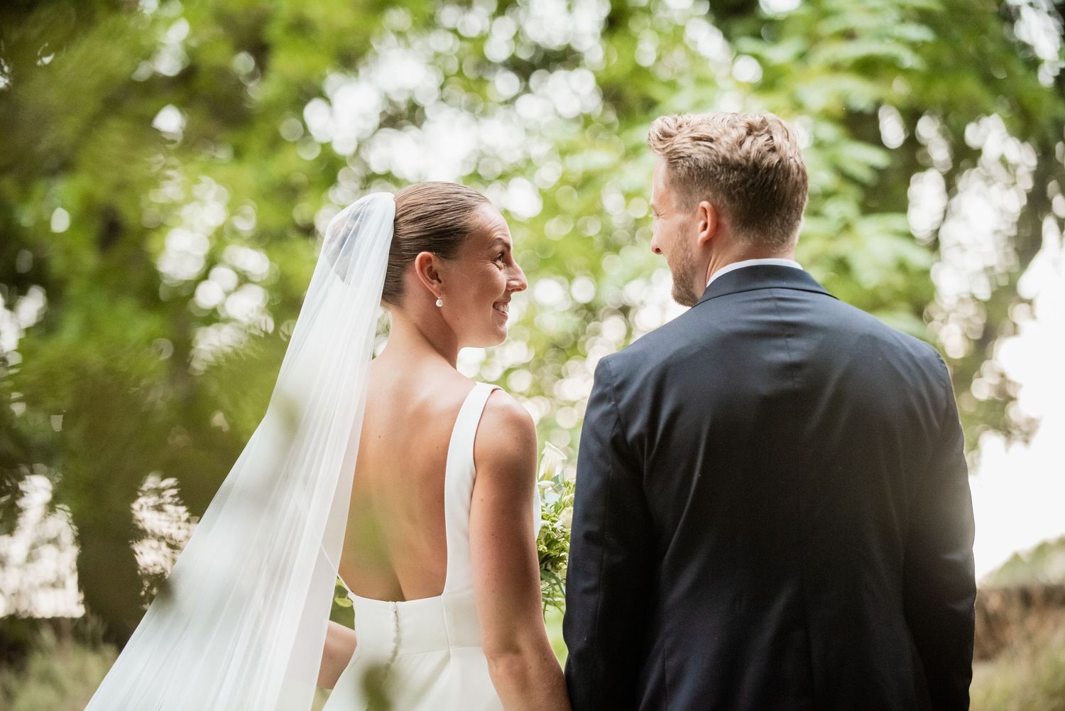 A bride in a white gown and veil holds hands with a groom in a navy suit, standing outdoors surrounded by lush green trees.