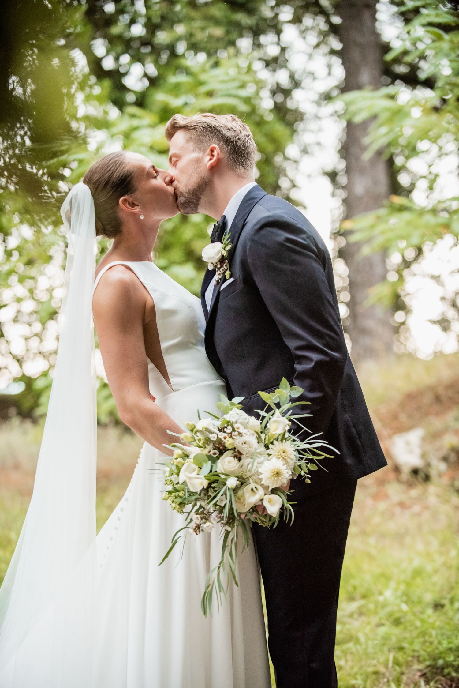 Bride and groom sharing a kiss outdoors, bride holding a white and greenery bouquet, groom in a navy suit.