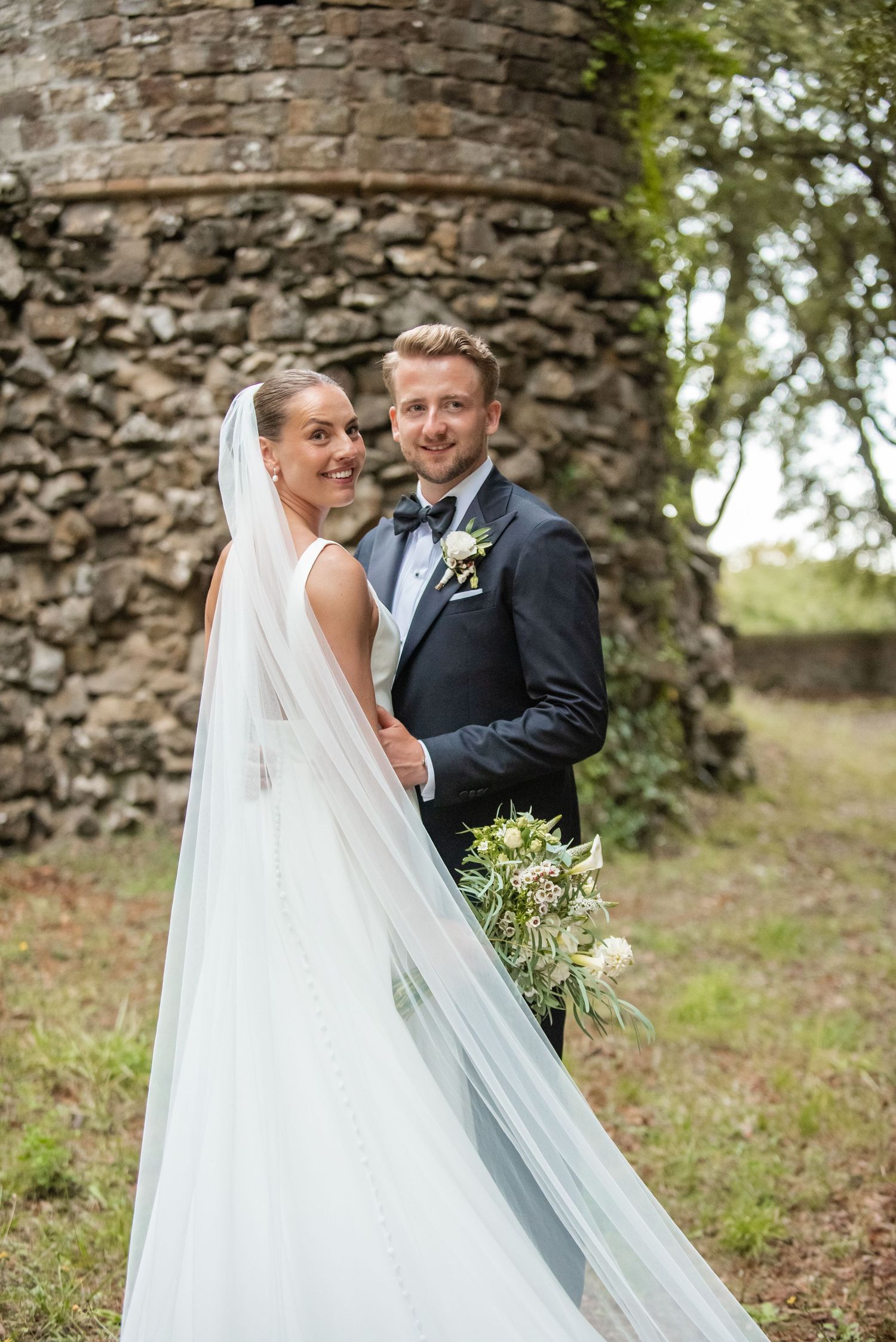 Bride in white gown with veil and groom in navy tuxedo pose together near a stone tower outdoors.
