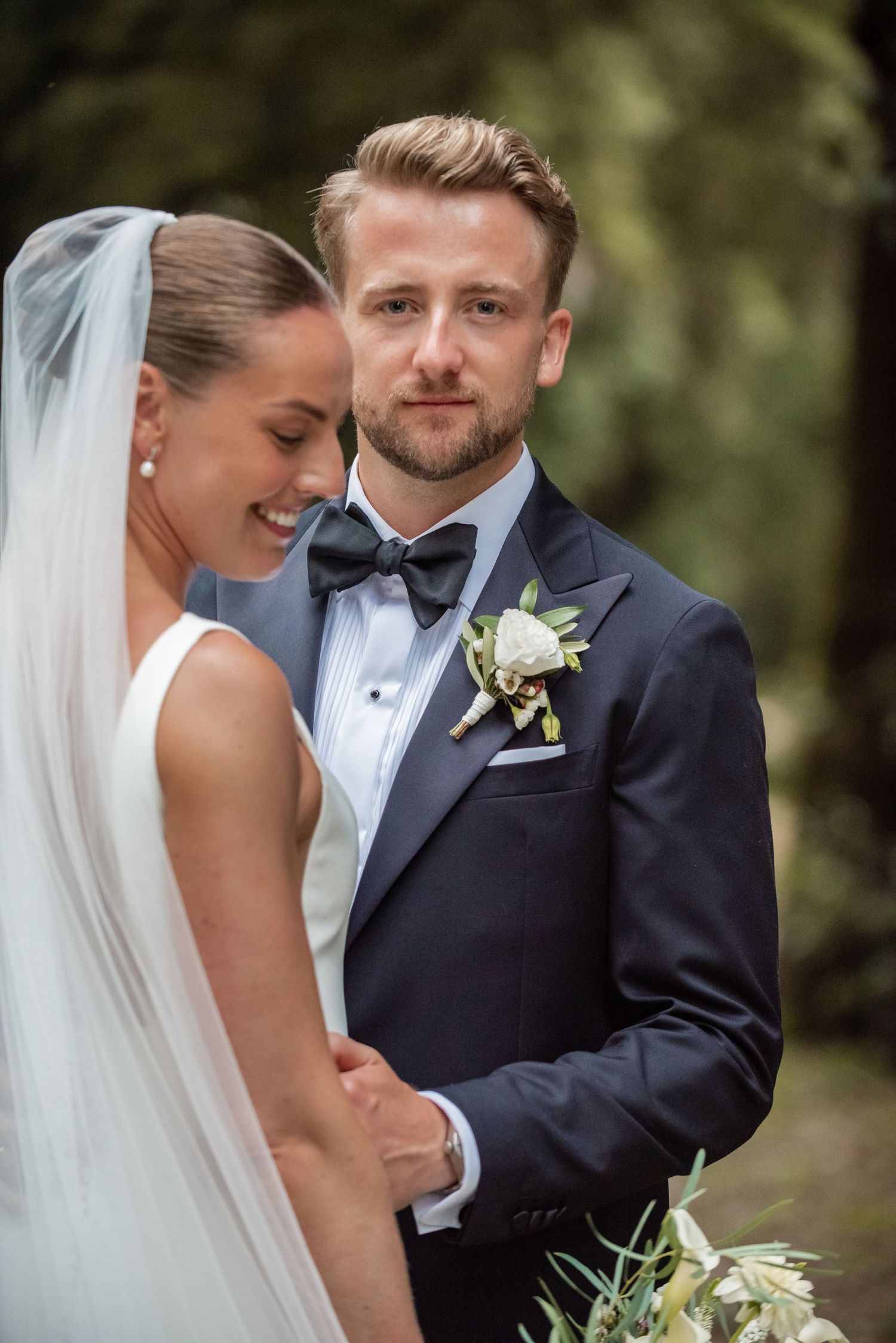 A bride in a white gown and veil leans into a groom wearing a navy tuxedo with a floral boutonniere outdoors.