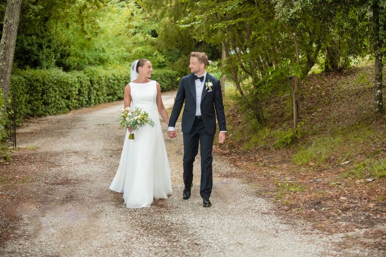 Bride and groom walking hand in hand on a tree-lined gravel path, bride in white gown holding bouquet.