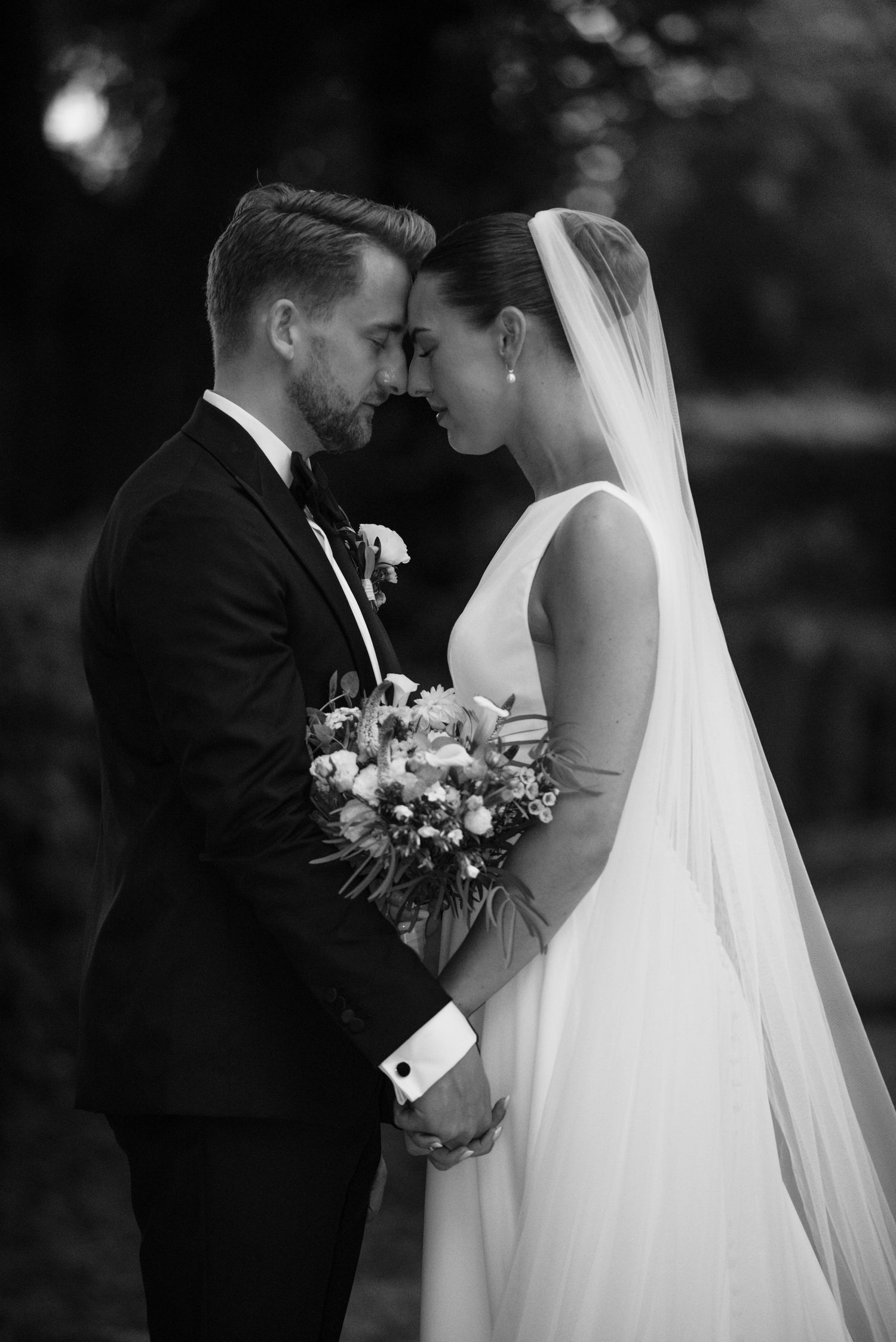 Black and white wedding photo of bride and groom touching foreheads, bride holding bouquet in elegant gown with veil.
