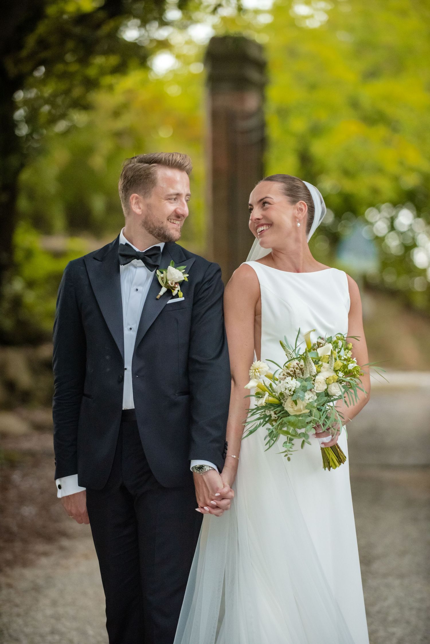 A smiling bride holding a white bouquet and groom in black tuxedo hold hands outdoors in a lush green garden setting.