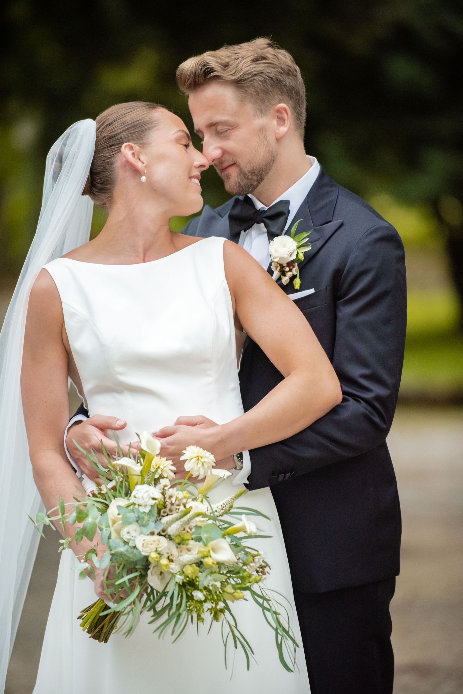 A bride and groom share a romantic moment, nearly kissing, as she holds a white and green bouquet outdoors.
