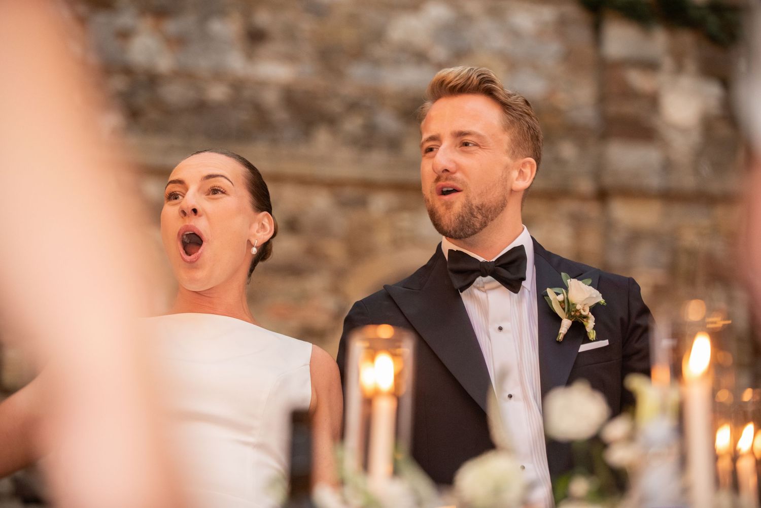 Bride and groom singing joyfully at candlelit wedding reception, bride in white gown, groom in navy tuxedo with boutonniere.