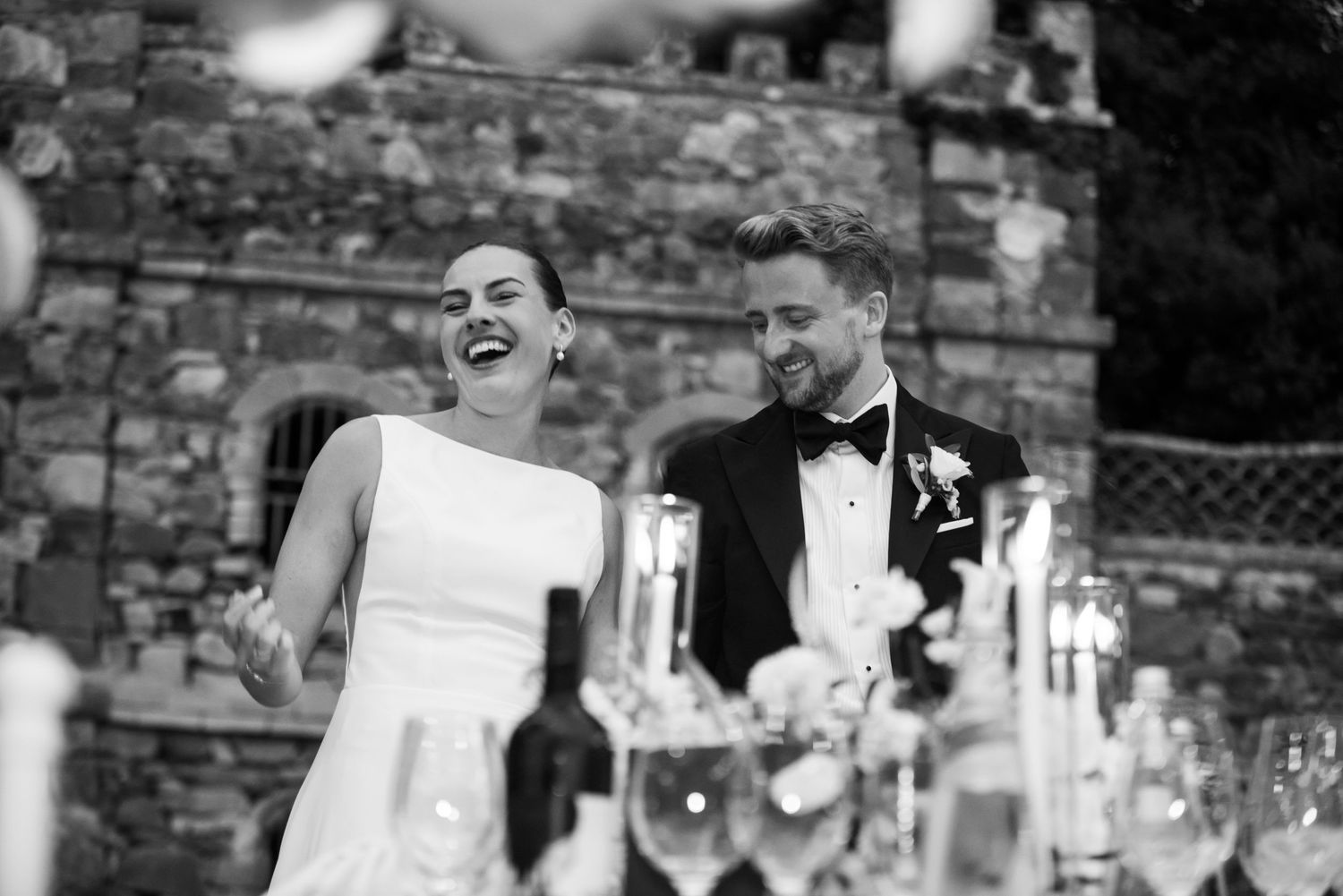 Black and white photo of a laughing bride and groom in formal attire at an elegant outdoor wedding reception.