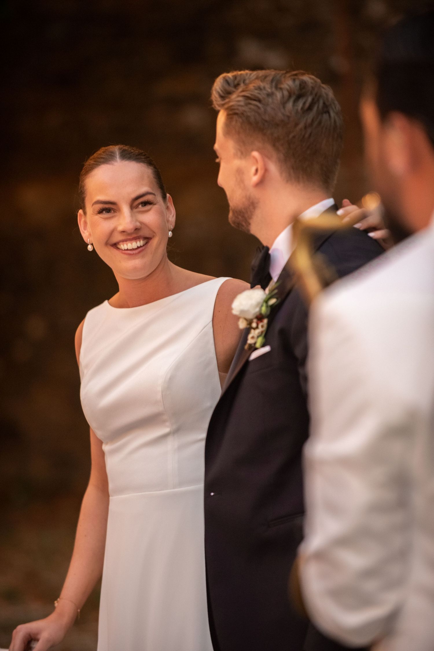Bride in white gown smiling joyfully at groom in black tuxedo during outdoor wedding ceremony.