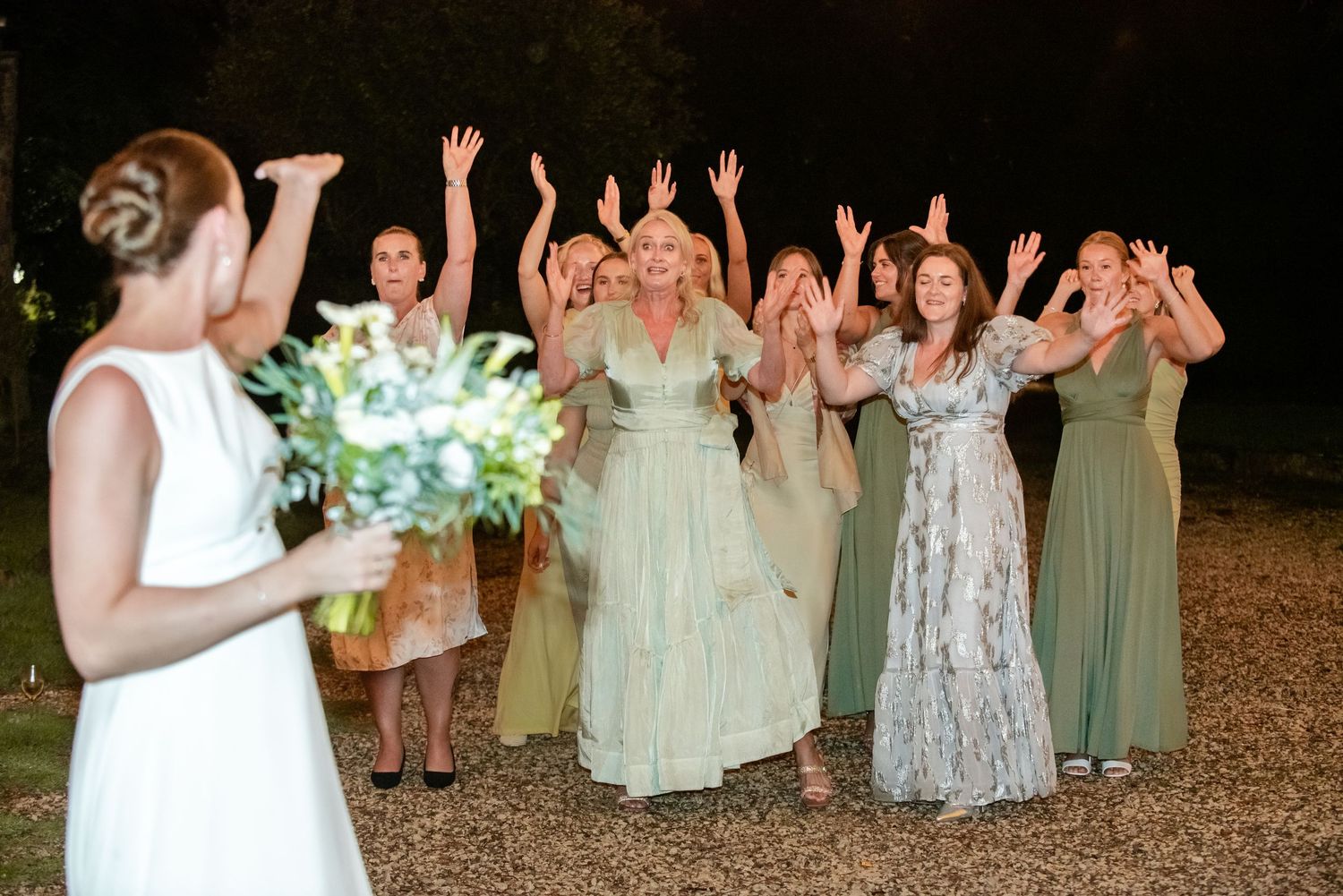 Bride tossing bouquet to excited bridesmaids in green dresses at outdoor evening wedding celebration.