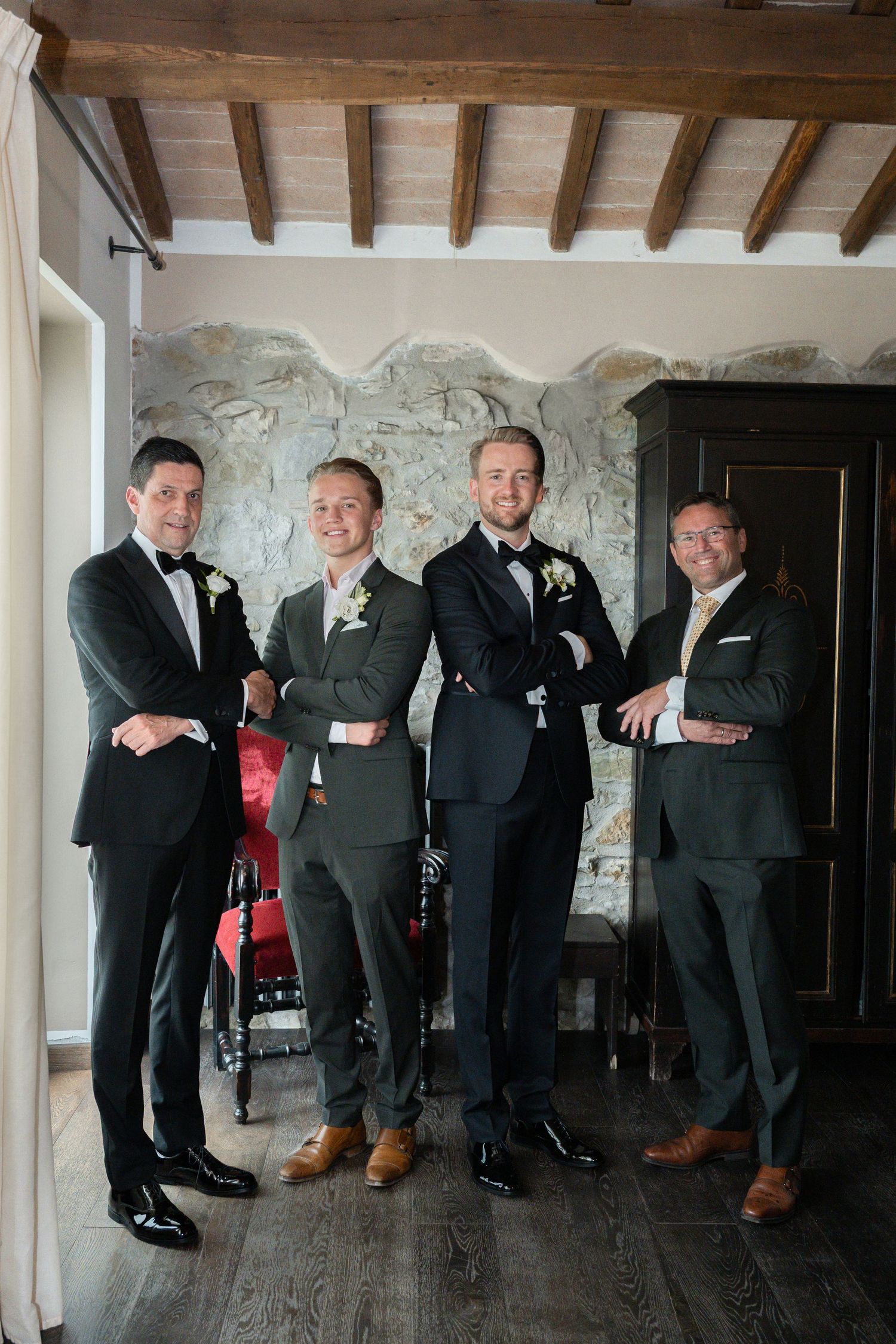 Four groomsmen in dark suits with boutonnieres pose with crossed arms in a rustic stone-walled venue.