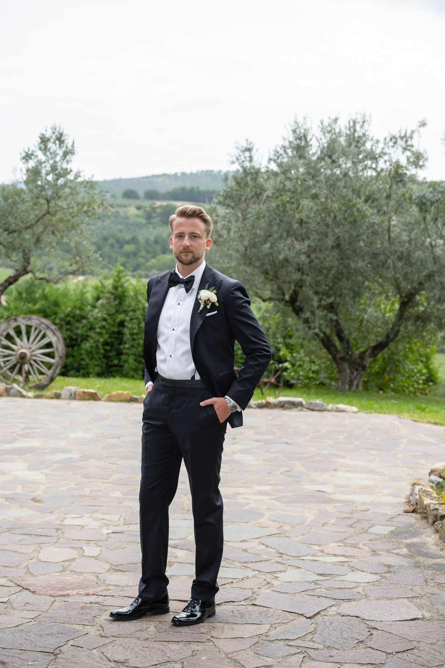 Groom in black tuxedo with boutonniere stands confidently on stone patio with olive trees and hills in background.