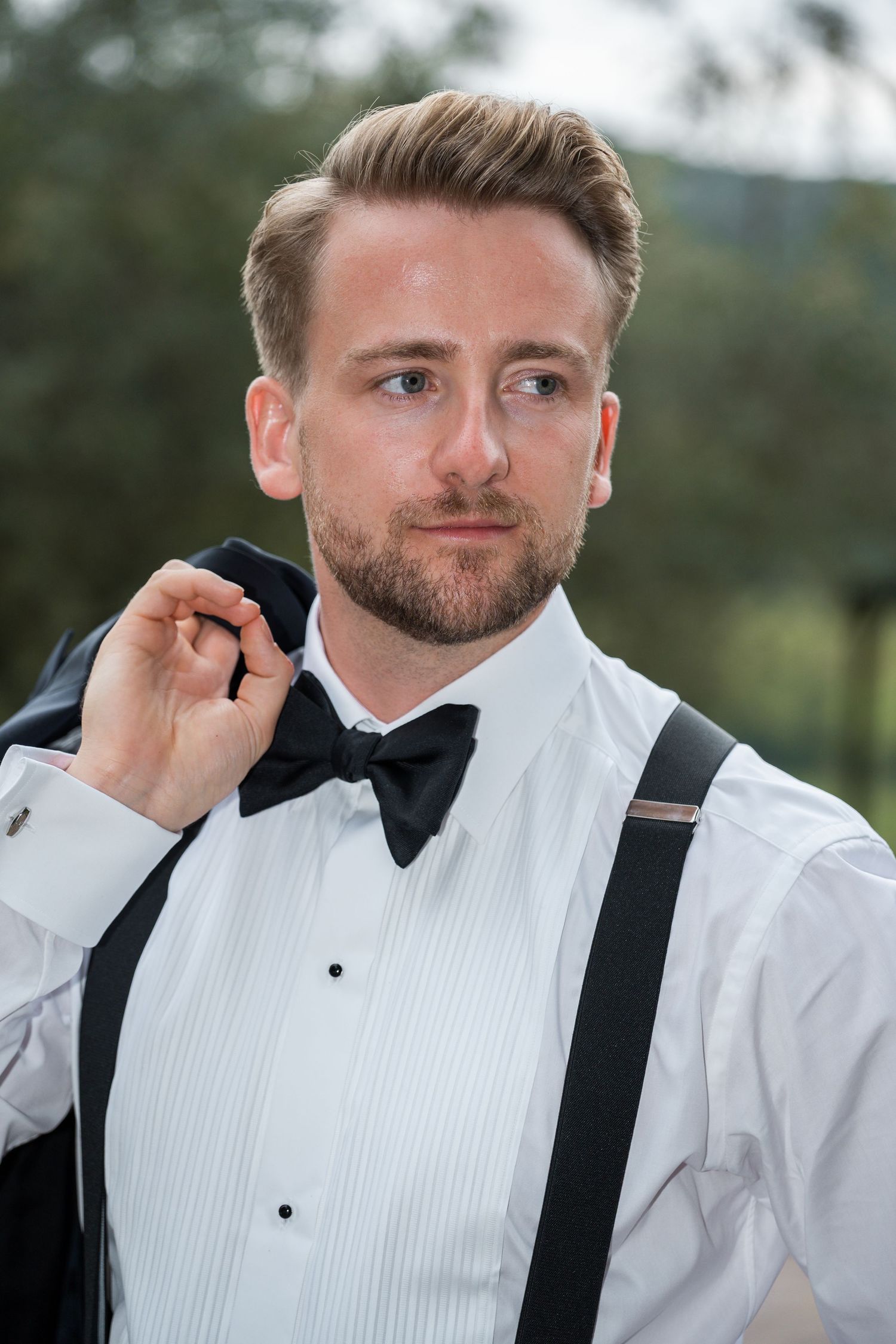A dapper groom in a white tuxedo shirt, black bow tie, and suspenders holds his jacket over his shoulder outdoors.