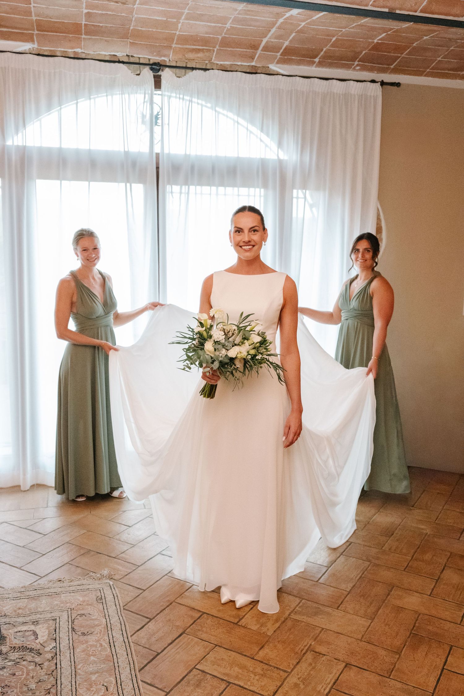 Bride in white gown holds bouquet while two bridesmaids in sage green dresses hold her train indoors.