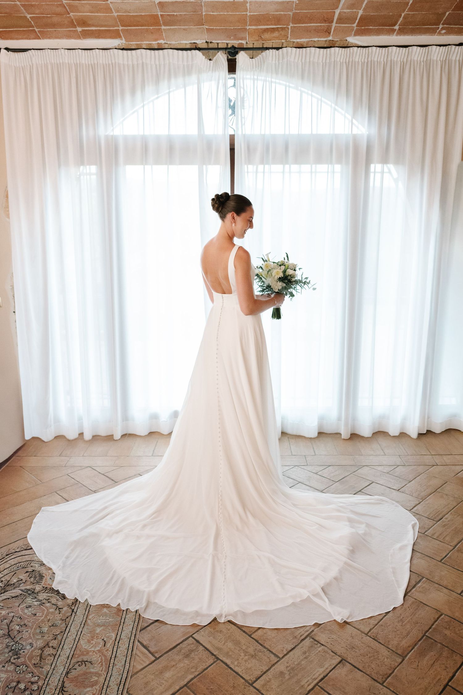 Bride in elegant white backless wedding gown with long train holds bouquet, standing before arched window with sheer curtains.