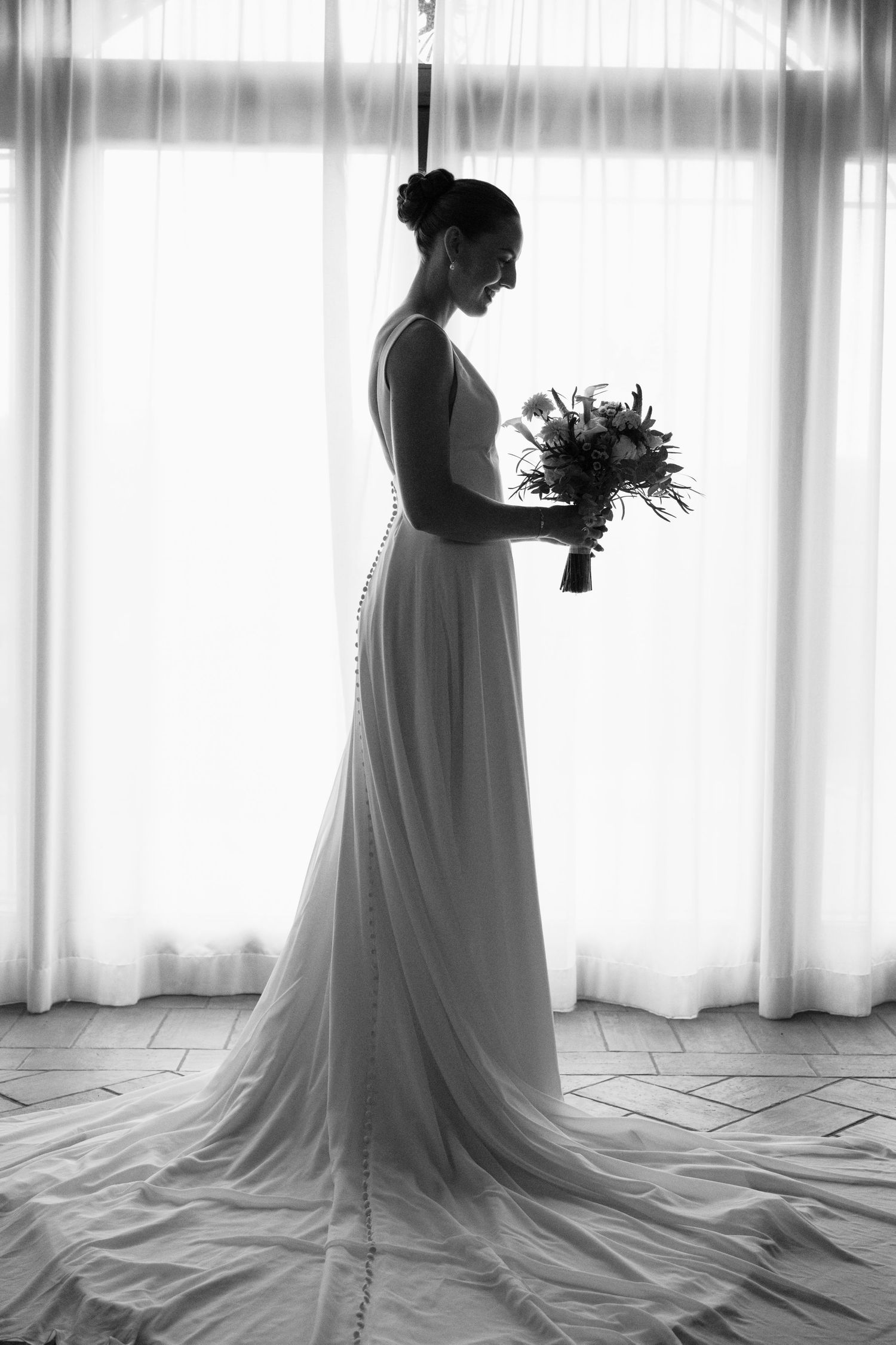 Black and white photo of a bride in a flowing gown holding a bouquet, standing gracefully before a bright window.