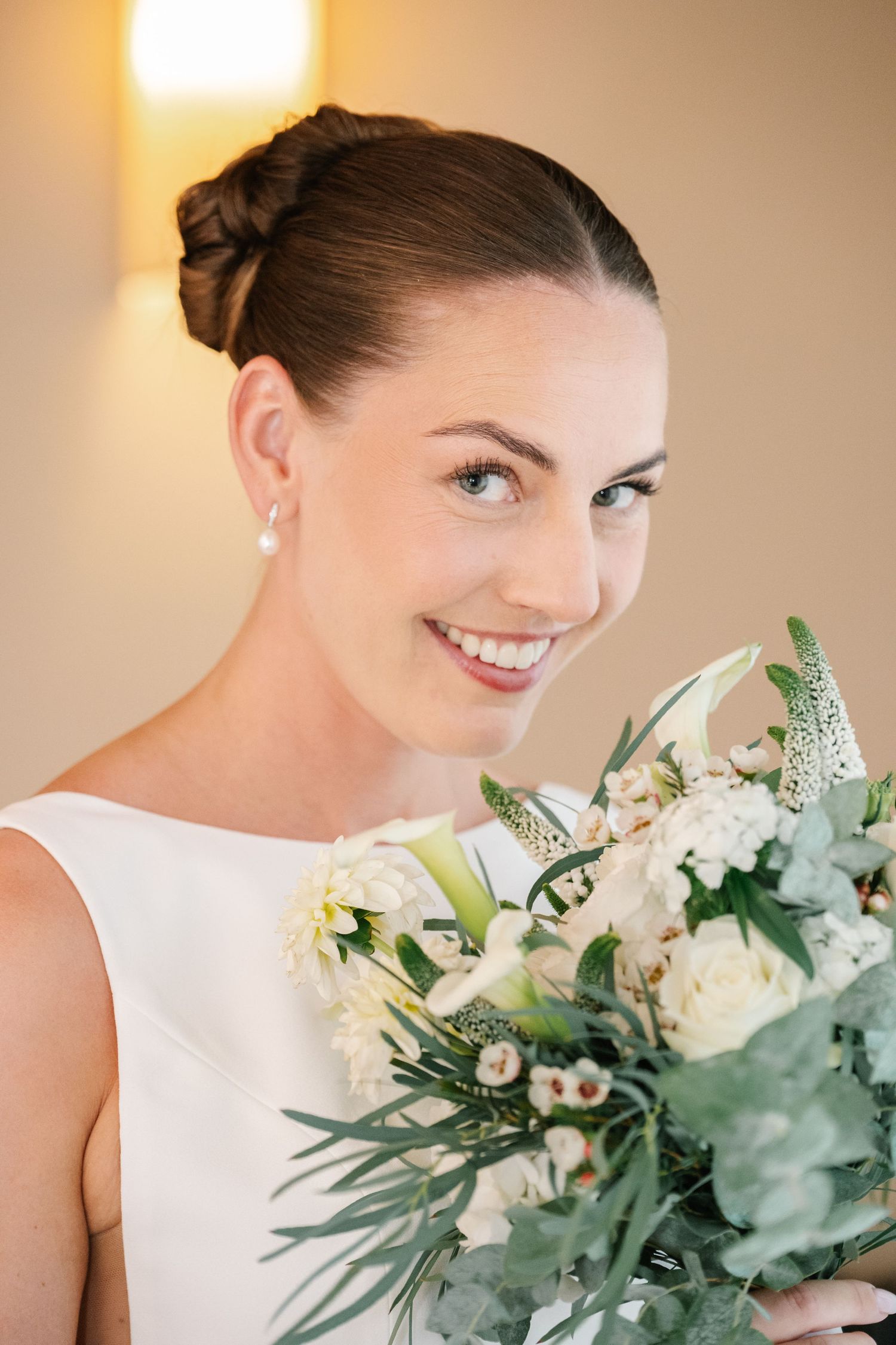 Smiling bride in white dress holding elegant bouquet of white roses and greenery, with updo hairstyle and pearl earrings.